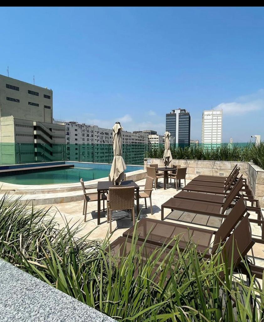 A rooftop terrace features a swimming pool surrounded by sun loungers and shaded umbrellas. Lush green grass adds a natural element to the space, with city buildings visible in the background under a clear blue sky.