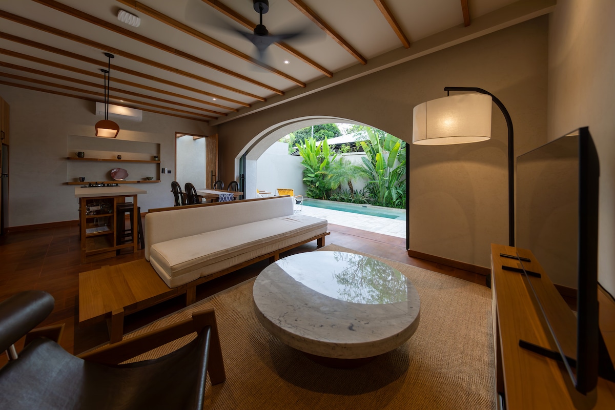 An open-concept living area features a comfortable sofa and a round marble coffee table. Natural light pours in through arched doors, revealing a view of the private pool and tropical greenery. A wooden dining table and stylish ceiling fan complement the serene atmosphere.