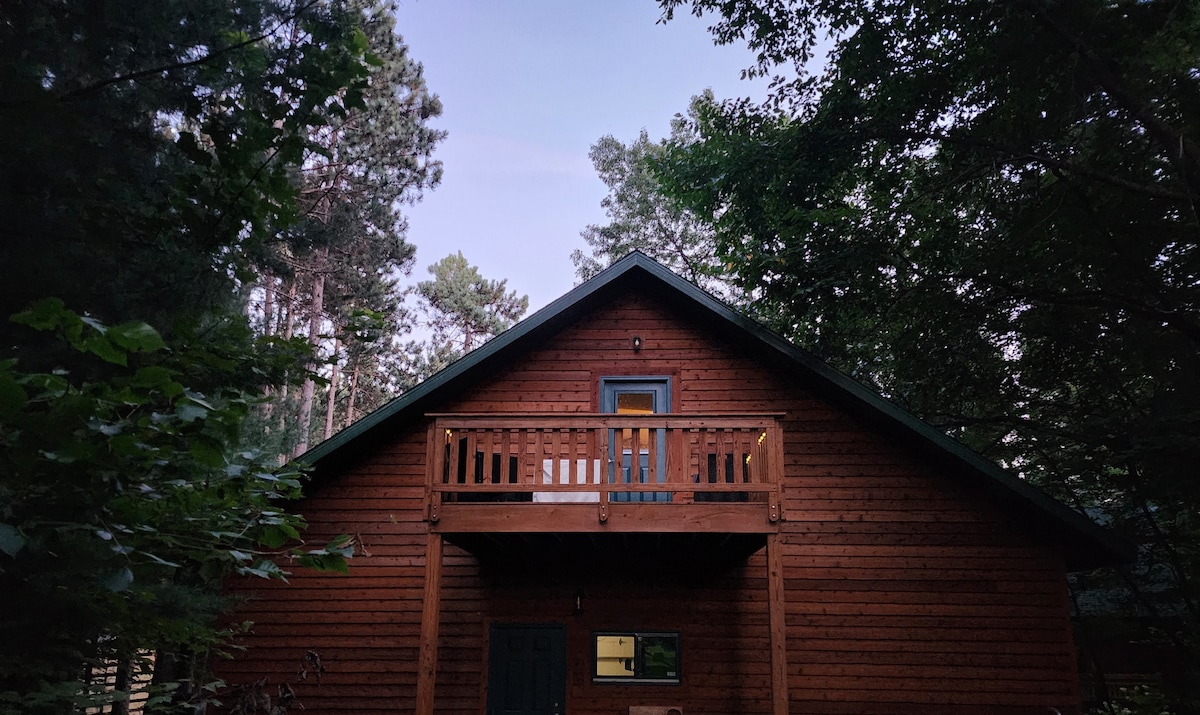 A charming wooden cabin is framed by tall trees, showcasing a spacious balcony with wooden railings. The front entrance is visible, complemented by an inviting warm glow from a window, creating a serene atmosphere during twilight.