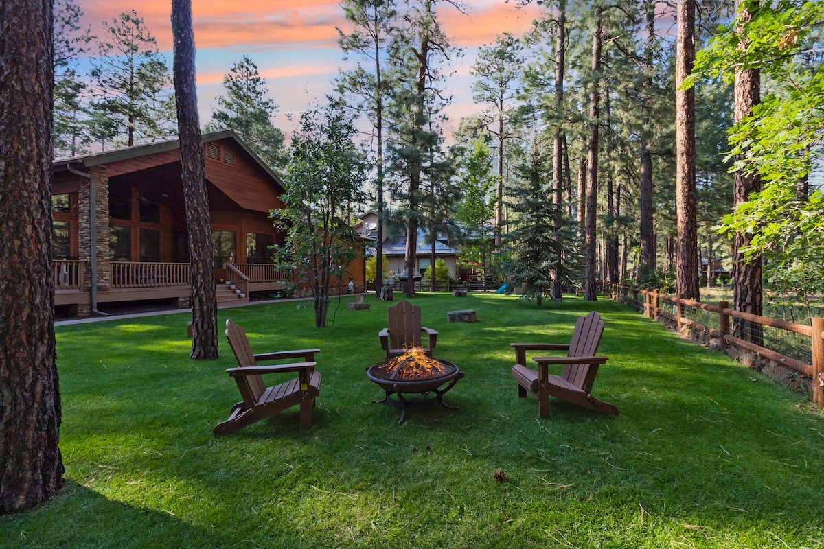 A serene outdoor space features comfortable Adirondack chairs arranged around a fire pit, surrounded by lush green grass and towering ponderosa pine trees. The inviting landscape encourages relaxation and connection with nature, enhanced by a vibrant sunset sky in the background.