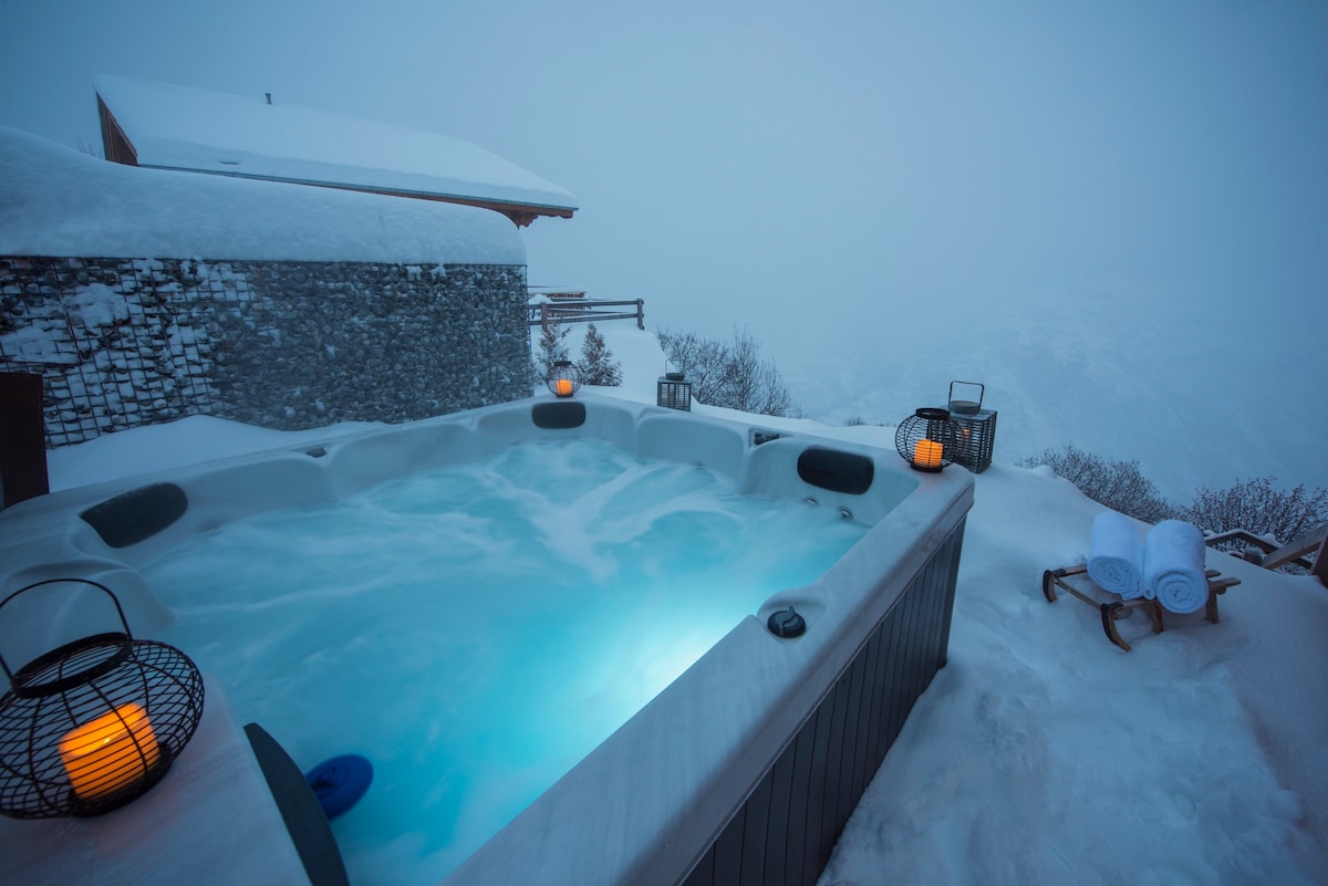 An outdoor jacuzzi is surrounded by fresh snow, creating a serene winter oasis. Soft blue water contrasts against the white landscape, while nearby lanterns provide gentle illumination. Towels are neatly arranged on a ledge, enhancing the inviting atmosphere for relaxation after a day in the snow.