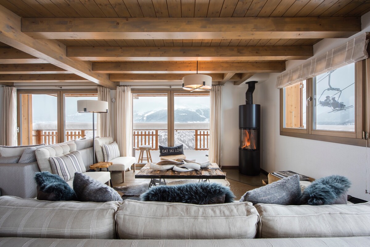 A spacious living area features comfortable seating arranged around a central coffee table. Large windows allow natural light to fill the room while offering views of the snowy landscape. A modern fireplace adds warmth to the atmosphere, complemented by wooden beams overhead.