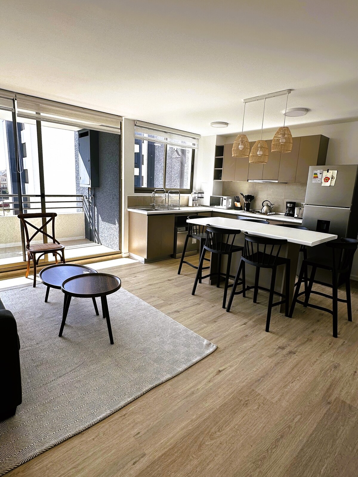 A well-lit kitchen and living area features a countertop with seating for six. Natural light fills the space through floor-to-ceiling windows, with a small round table nearby. Wooden accents and a light rug contribute to an inviting atmosphere.