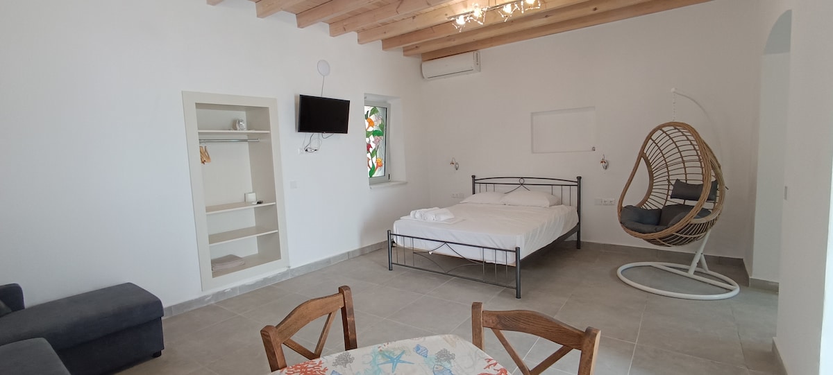 A spacious room features a comfortable bed framed in black metal, complemented by a modern hanging chair. A small dining area is present with wooden chairs around a table. Natural light enters through a colorful stained glass window, enhancing the bright atmosphere.