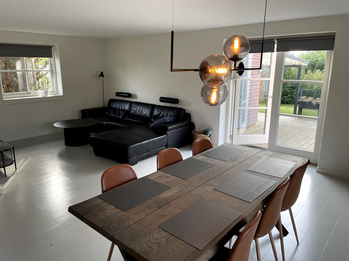 A modern living space features a large dining table surrounded by chairs. A black sectional sofa is positioned nearby, while light streams in from the glass doors that open to the garden. Clean lines and neutral tones create a spacious atmosphere.