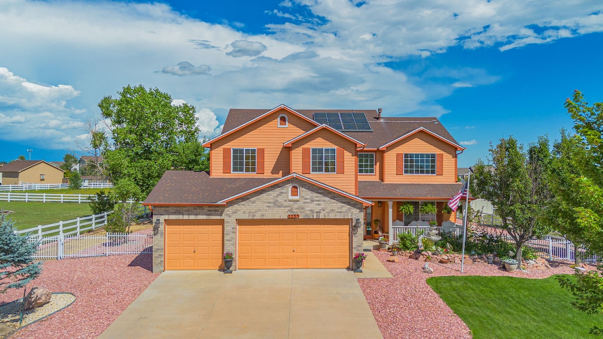 The exterior of a spacious two-story home is showcased, featuring a mix of orange and stone siding. Solar panels are visible on the roof. A wide driveway leads to a double garage. Lush greenery and decorative landscaping surround the property, with a white fence partially visible.