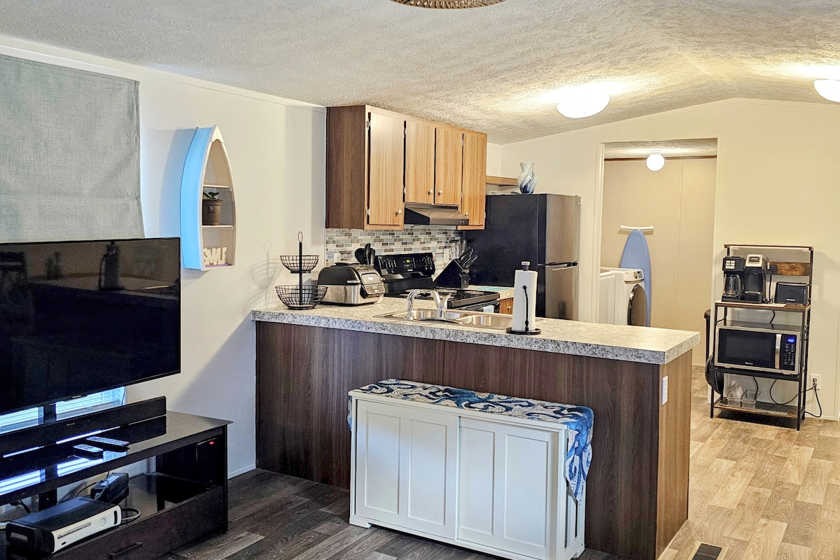 A modern kitchen area is portrayed, featuring wooden cabinetry and a dark countertop. An adjustable bench is situated near the front, with various kitchen appliances visible in the background. The open layout connects to a cozy living space, ensuring ample room for movement.
