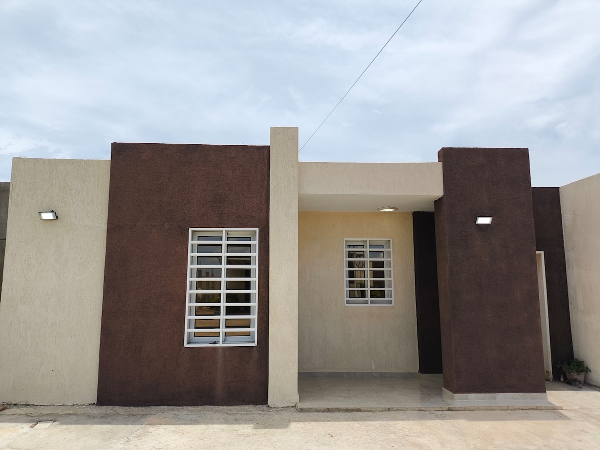 An exterior view of a modern residential building featuring a combination of textured brown and light-colored walls. Large windows with horizontal slats are visible, letting in natural light. The entrance is framed by minimalistic lighting fixtures, contributing to a clean and welcoming design.