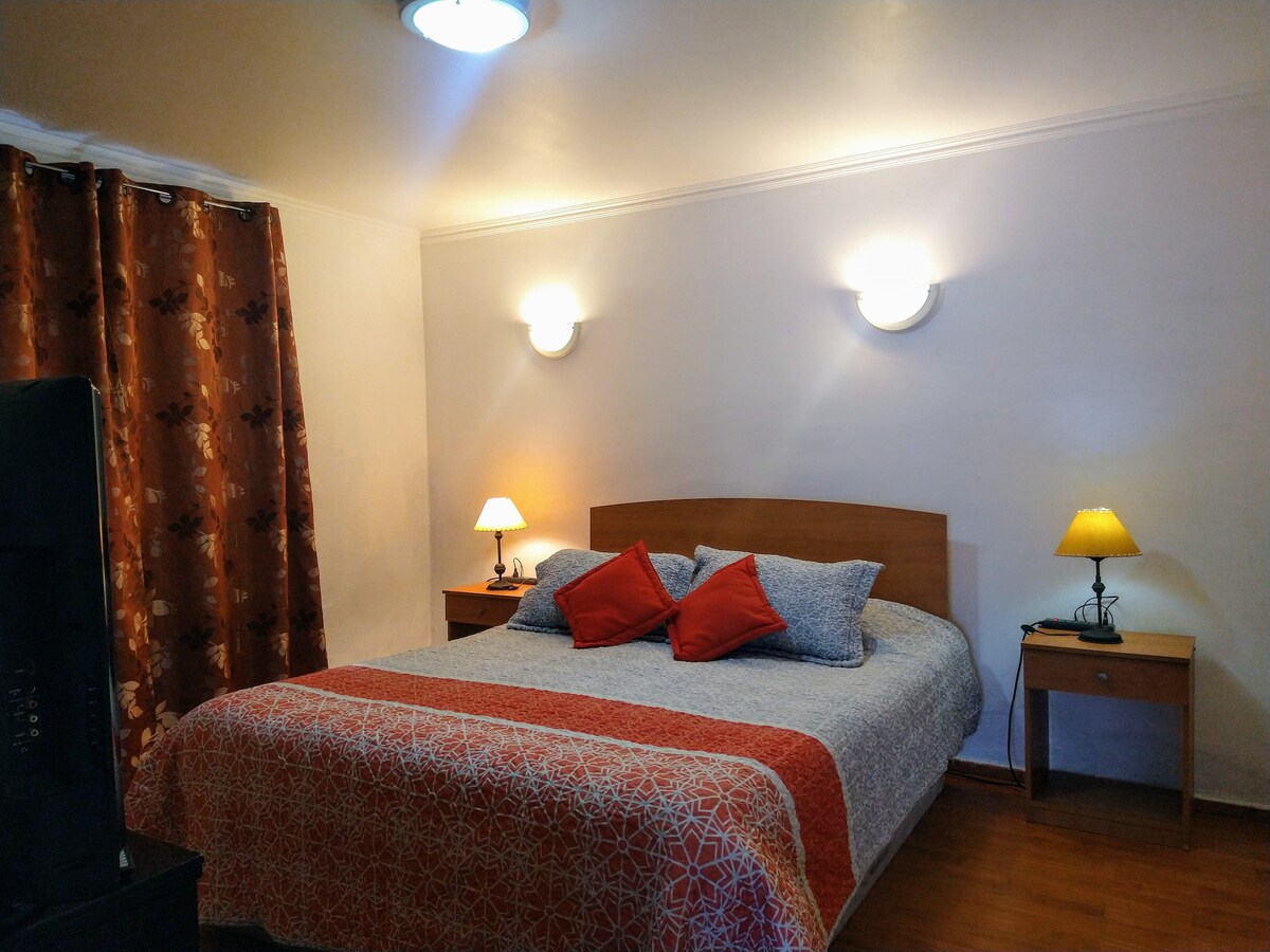 A well-appointed sleeping area features a spacious bed with decorative pillows and a patterned bedspread. Two bedside tables are positioned on either side, each with a lamp. Soft lighting from wall fixtures creates a warm atmosphere, complemented by curtain-covered windows.