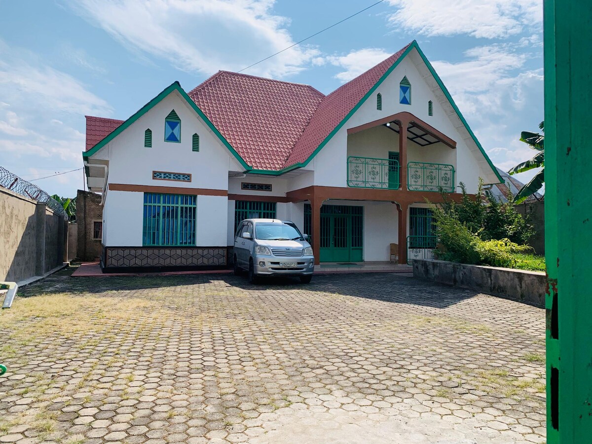 A spacious house with a distinctive architectural style is set in a well-maintained outdoor area. A vehicle is parked in the stone-paved driveway, and the exterior features large windows with blue accents. Lush greenery is visible in the surrounding landscape.