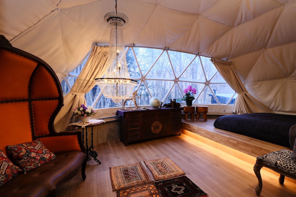 A well-appointed dome-shaped room features a striking antique chandelier set against a backdrop of large windows. An elegant seating area with colorful cushions complements the wooden furnishings. Natural light fills the space, enhancing the tranquil ambiance created by the surrounding forest.