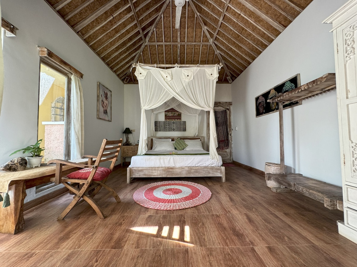 A spacious double room features a four-poster bed draped with white fabric, complemented by a rustic wooden table and chair. Natural light enters through an oversized window. Additionally, a circular rug adds warmth to the wooden floor, and art pieces adorn the walls.