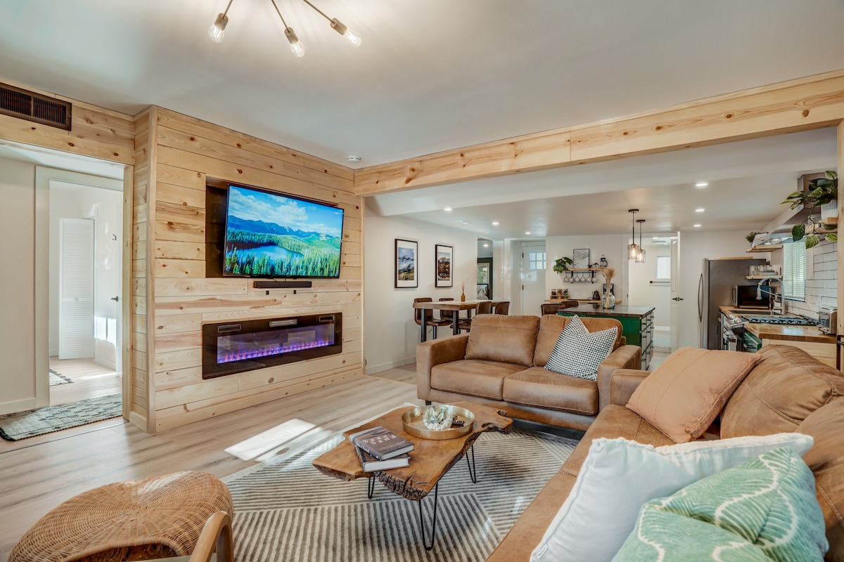 An open living area features natural wood paneling, a cozy brown sofa, and a unique coffee table. A large flat-screen Smart TV is mounted on the wall, with ambient lighting beneath a sleek electric fireplace. The kitchen and dining area are visible in the background.