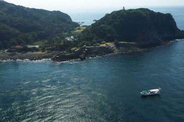 Aerial view of a serene coastal landscape where the turquoise ocean meets rocky shorelines. A small boat is seen traversing the water near a green hill, accented by trees and natural vegetation. The coastline is dotted with homes, harmonizing with the peaceful environment.