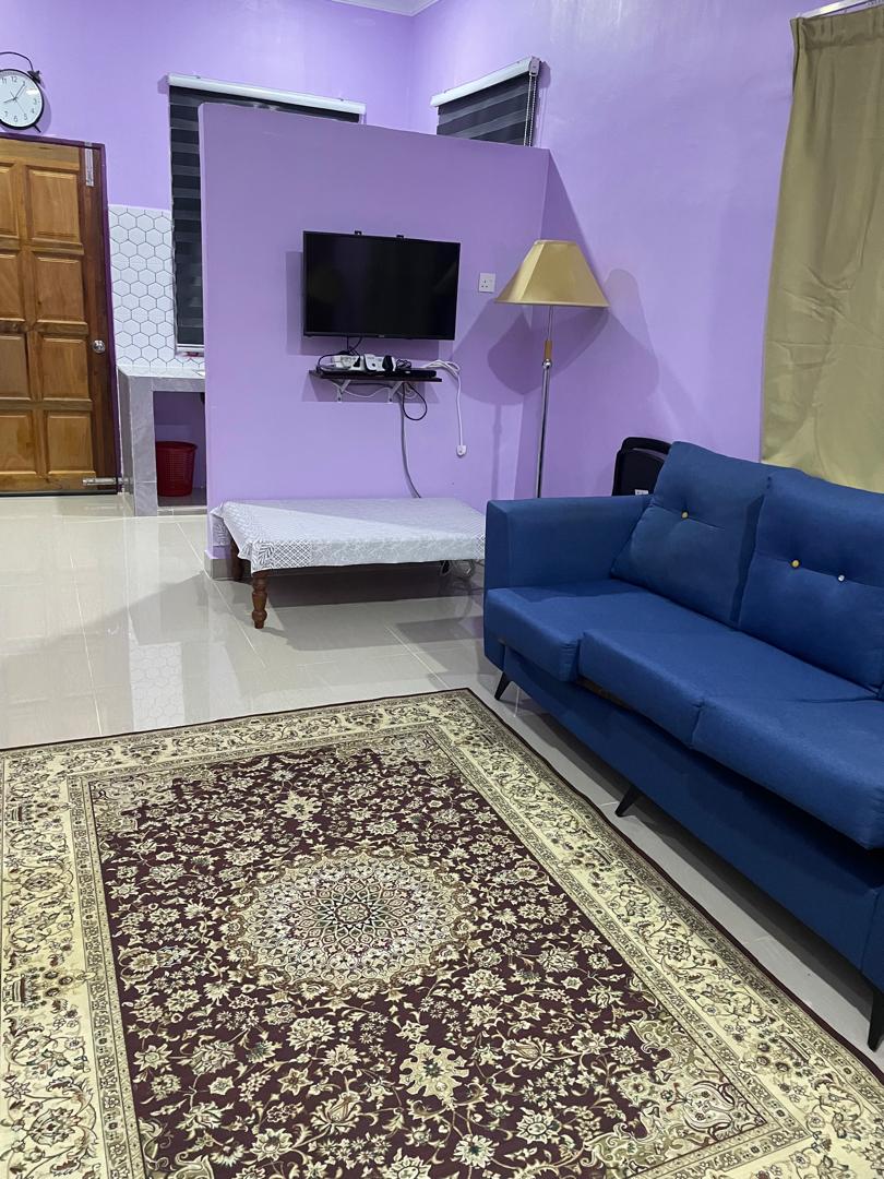 A comfortable seating area is featured with a blue sofa positioned on an intricate area rug. A wall-mounted television is visible, accompanied by a standing lamp. The room is painted in a soft purple hue, complemented by two windows with dark blinds.