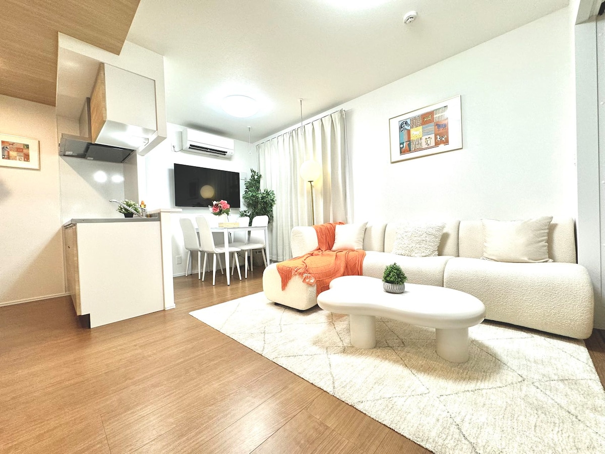 A spacious living area showcases a contemporary design with light wood flooring. A comfortable sofa is adorned with an orange throw blanket. A round coffee table sits centrally, while a dining table is visible in the background, accompanied by four chairs. Natural light enters through sheer curtains.
