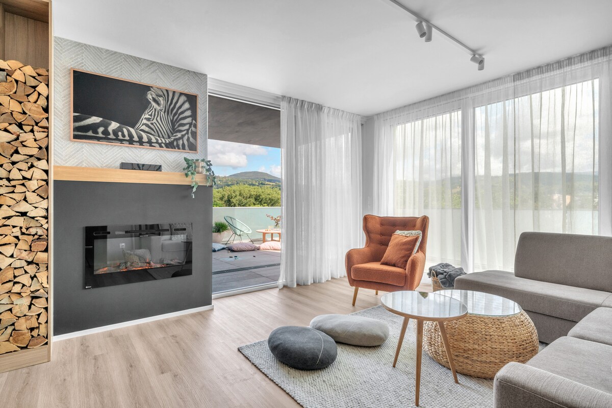 A bright living area features a modern gray sofa and an orange armchair, complemented by a small circular table. A cozy fireplace is integrated into the wall, and large windows provide a view of the outdoors, enhancing the natural light in the space.