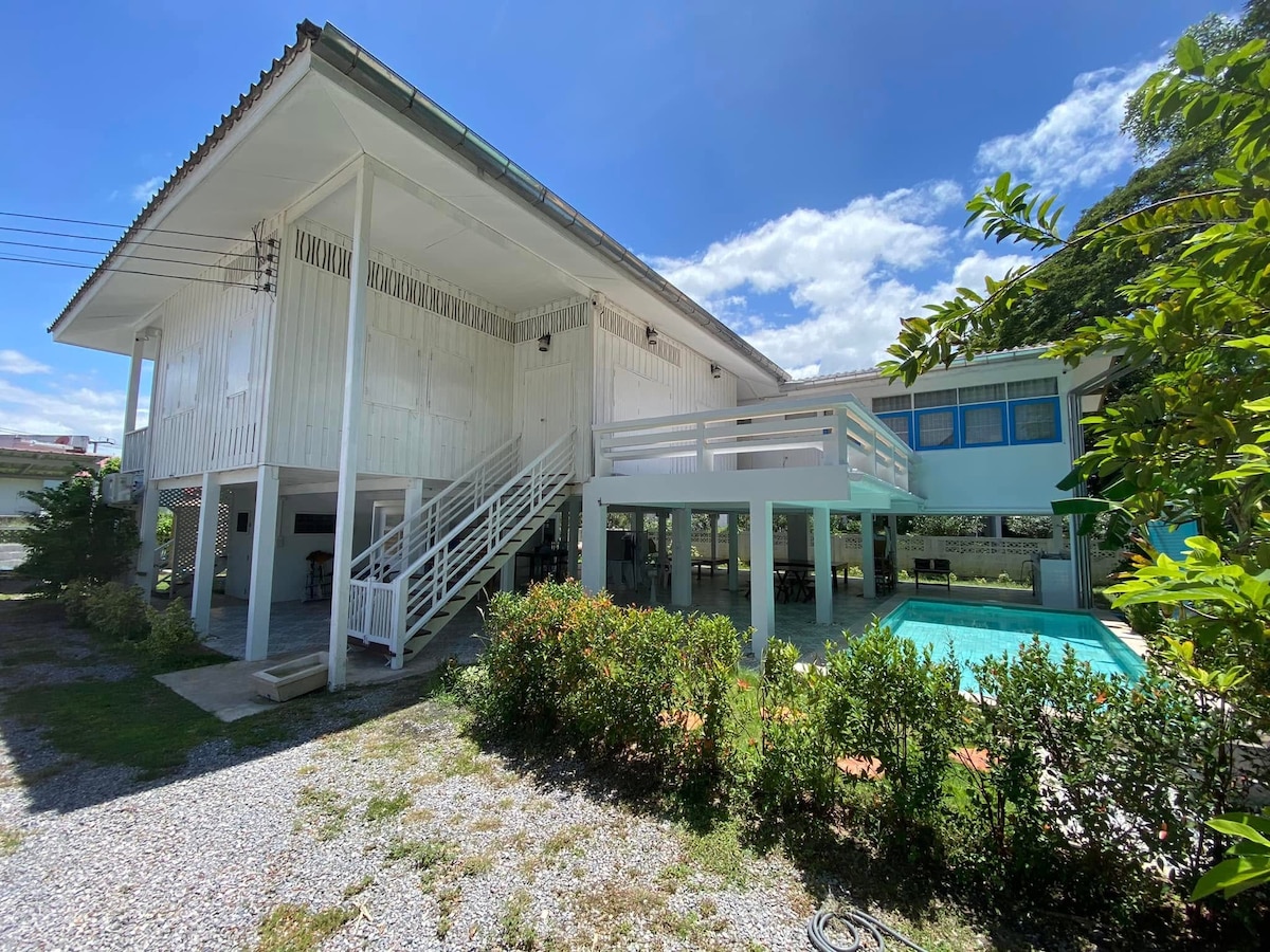 The exterior of the two-story white wooden house features a spacious area surrounded by lush greenery. A clear swimming pool is visible in front, complemented by a shaded terrace. Natural light illuminates the scene under a bright blue sky.