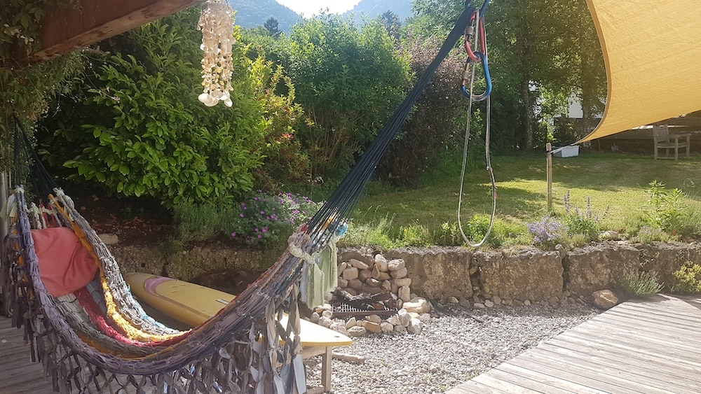 A colorful hammock is suspended under a sunshade, surrounded by lush greenery. Nearby, a stone wall frames a landscaped garden, featuring various plants and flowers. A wooden terrace is partially visible, with two surfboards resting against it, inviting relaxation in the peaceful outdoor space.