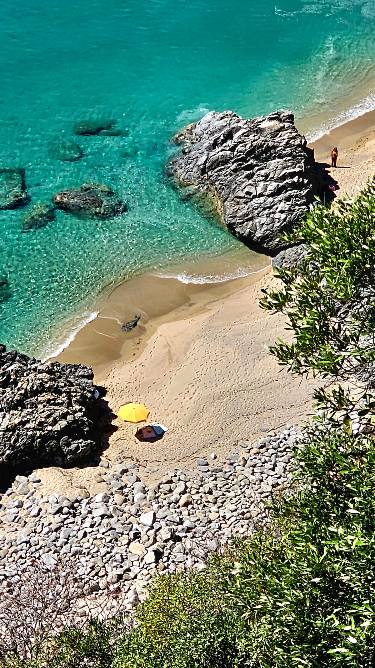 A sandy beach stretches along a clear turquoise coastline, bordered by rocky formations. Two beach umbrellas are positioned on the shore, while a few individuals are seen at a distance. The gentle waves wash onto the sand, enhancing the peaceful beach environment.