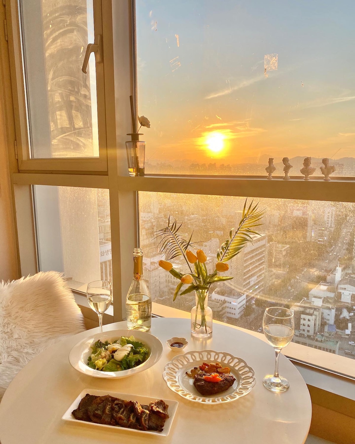 A dining table is set beside a large window, showcasing a sunset view over the city. A salad and a dish of meat are presented on elegant plates, accompanied by two glasses of white wine and a vase of fresh flowers. Soft lighting enhances the ambiance.