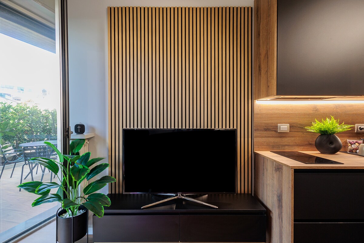A modern living area is depicted, featuring a wall-mounted television against a wooden slatted backdrop. A sleek cabinet below accommodates the TV, while a vibrant green plant adds a touch of nature. Large glass doors provide access to the private garden.