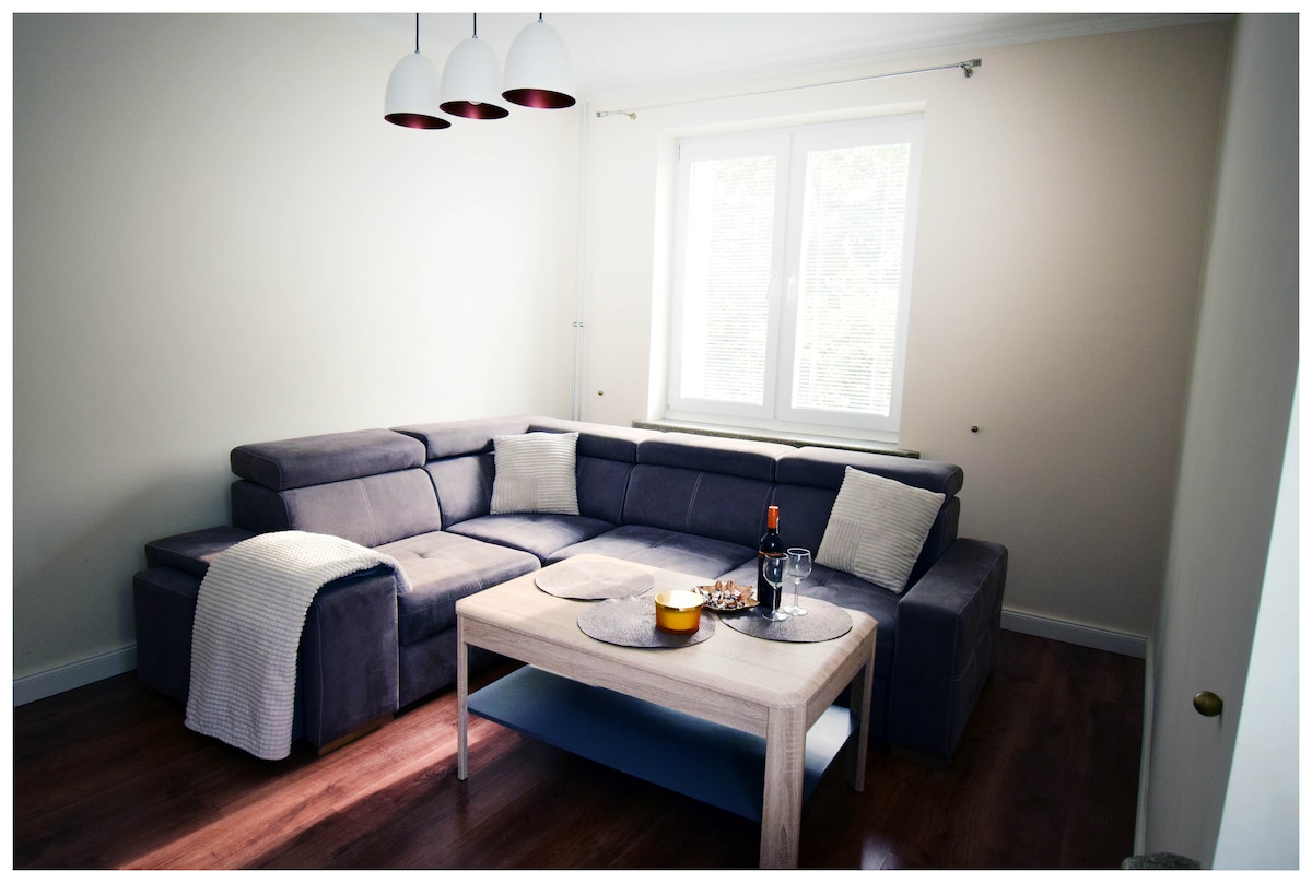 A comfortable gray sectional sofa is positioned in a well-lit living area. A light wooden coffee table holds decorative items and drinks. Natural light enters through a large window, complemented by three pendant lights above.
