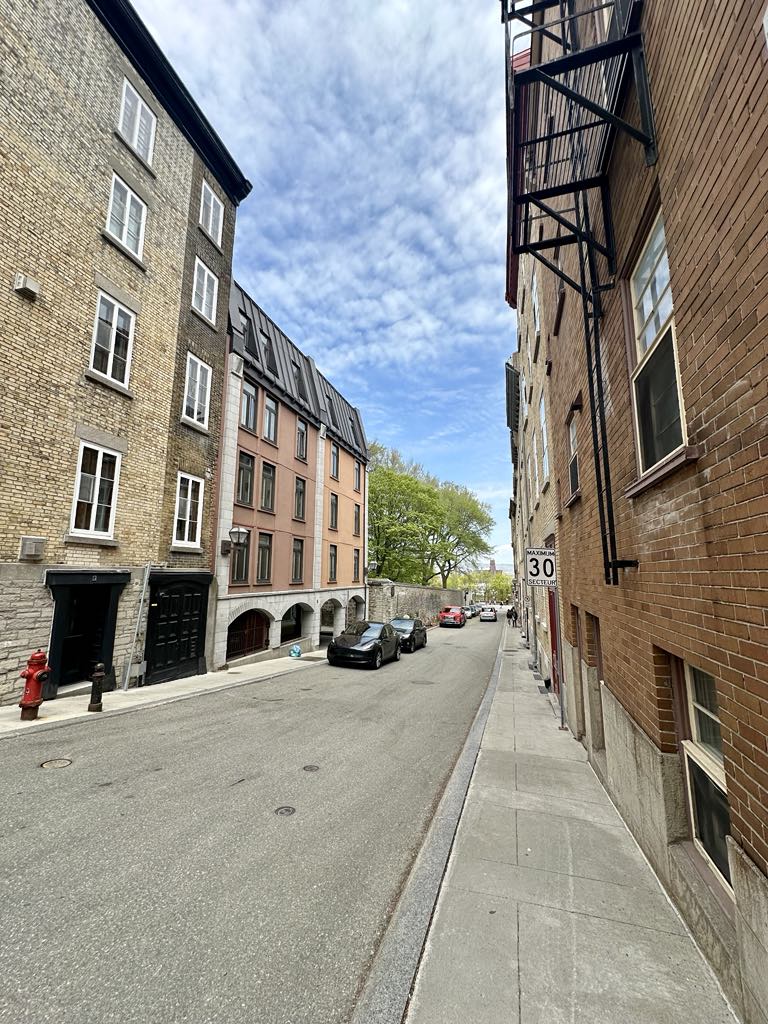A charming street scene in Quebec features a mix of historic brick buildings and modern architecture. Lush green trees line the street, while parked cars occupy the paved road. A clear blue sky with scattered clouds completes the serene setting.