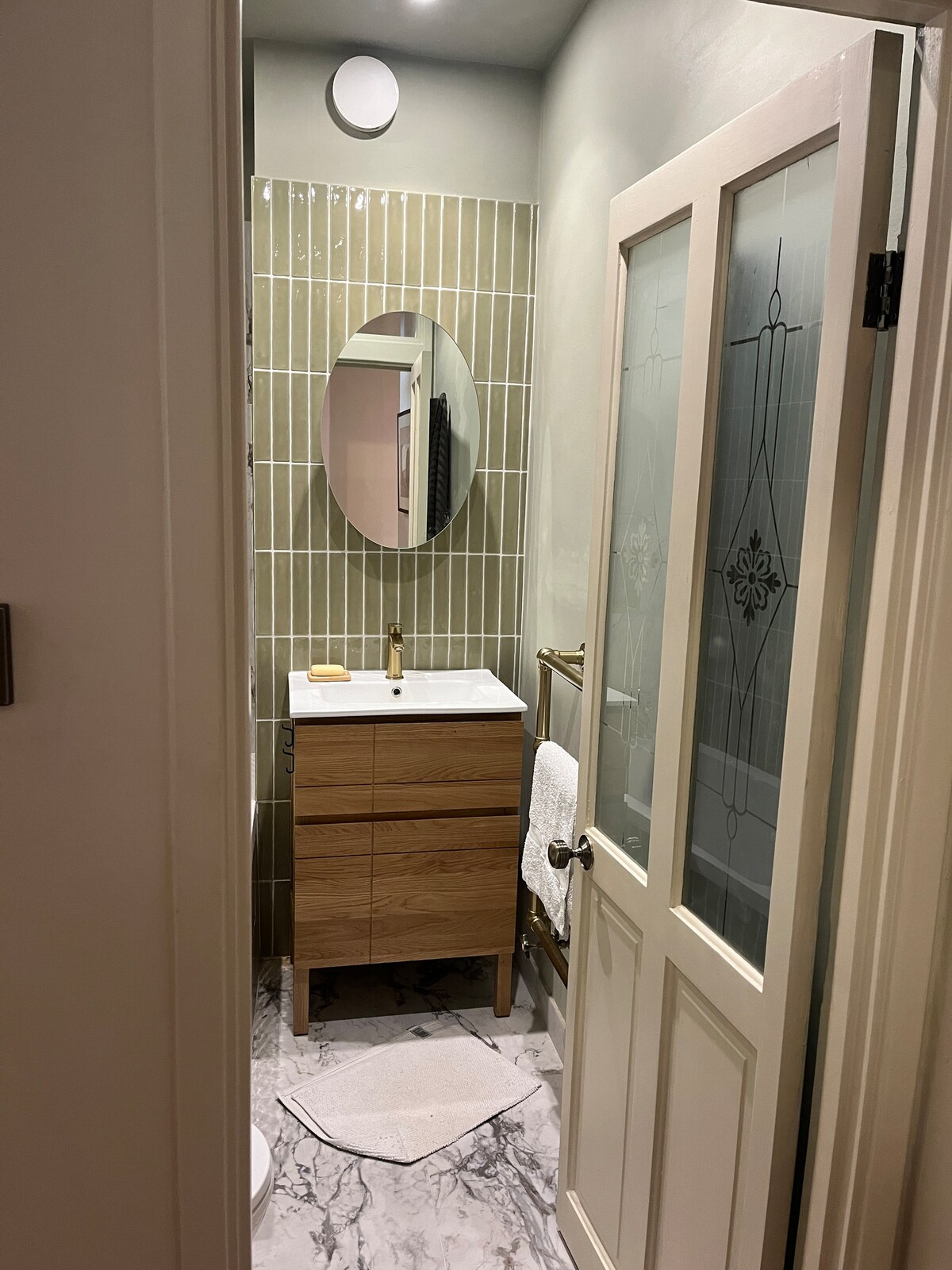 A compact bathroom features a wood-finished vanity with a white sink and gold faucet. Olive green walls and subtle tile detailing give a fresh look. An oval mirror hangs above the vanity, and a plush bath mat is placed on a marbled floor.