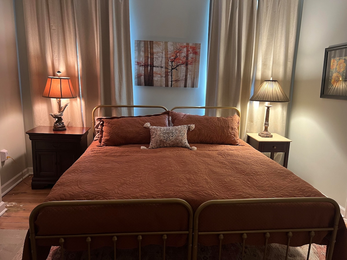 A spacious bed with an earthy-toned quilt and decorative pillows is framed by two lit bedside lamps. Soft curtains drape beside the bed, allowing gentle light to filter in. A piece of wall art featuring tree branches adds a touch of color to the room.