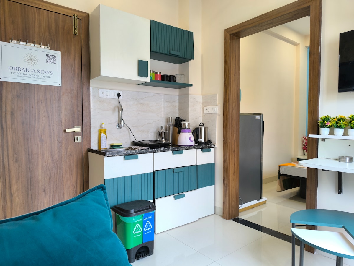 An open kitchen is depicted, featuring modern cabinetry in a teal and white color scheme. Appliances such as a kettle and coffee maker are positioned on the countertop. The area appears clean and organized, with a trash bin marked for recycling nearby.