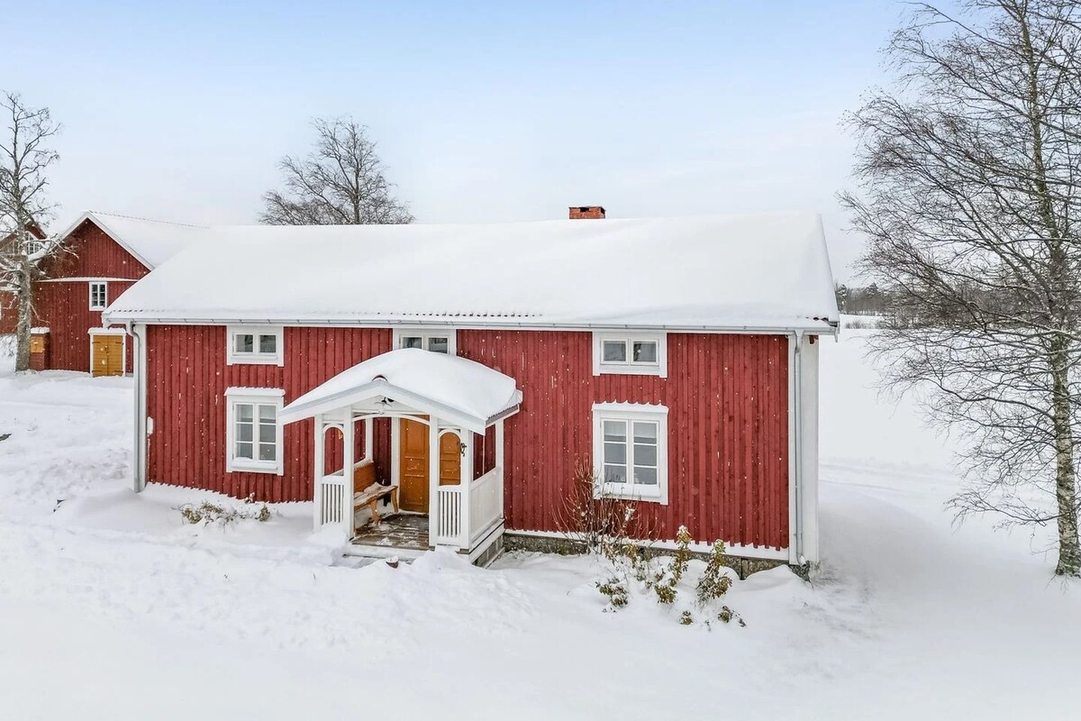 A charming red house with white trim is surrounded by a blanket of snow. The entrance features a covered porch with wooden doors, framed by snow-covered landscaping. Nearby trees stand bare against the winter sky, creating a serene, wintry scene.
