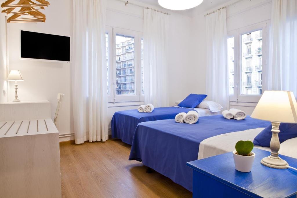 The room features two beds dressed in blue linens, with white throws. Natural light floods through multiple windows adorned with sheer white curtains. A wall-mounted TV is visible, and a desk with a vase of greenery adds a touch of decor. Towels are neatly arranged on the beds.
