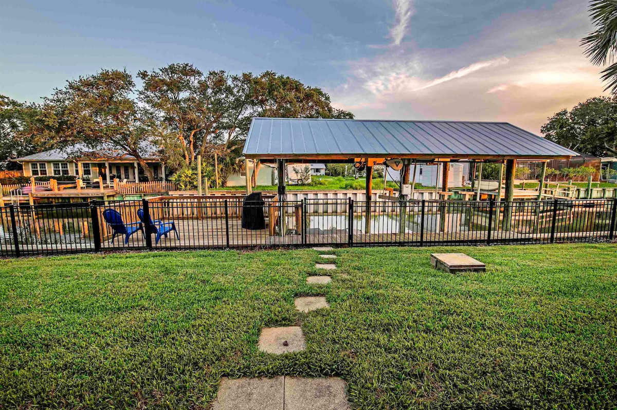 A serene backyard scene showcases a spacious lawn leading to a fenced outdoor area. A covered structure offers shade, with a grill visible nearby. The calm waters of the canal reflect the surrounding trees and sky, creating a peaceful environment for relaxation.