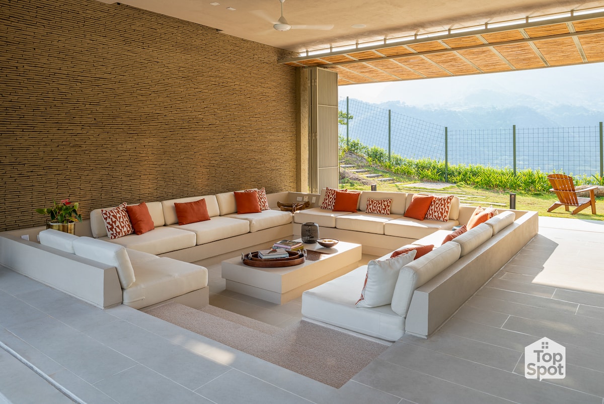 A modern outdoor lounge area features a spacious U-shaped sofa adorned with orange accent pillows. A low coffee table sits at the center, surrounded by a well-maintained lawn and panoramic mountain views visible through sliding glass doors.