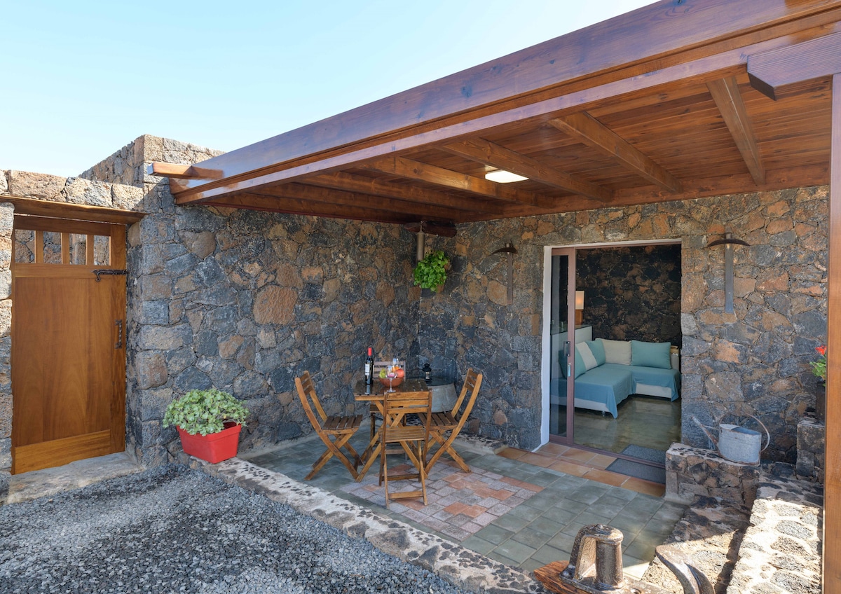 An outdoor seating area features a wooden table with two chairs under a shaded pergola. A vibrant potted plant adds a touch of green, while a stone wall enhances the rustic atmosphere. The interior view reveals a cozy living space with light-colored furnishings.