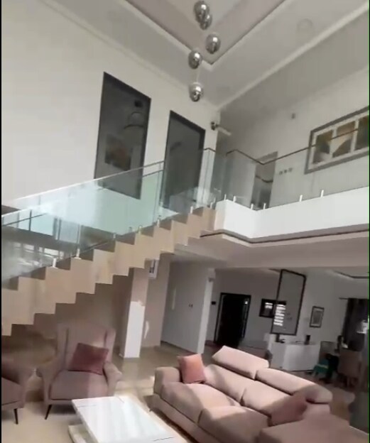 An open living area features modern furnishings, including a light-colored sofa and elegant armchairs. A glass staircase leads to an upper level, with large windows allowing natural light to fill the space. Decorative elements are visible on the walls, creating a contemporary atmosphere.