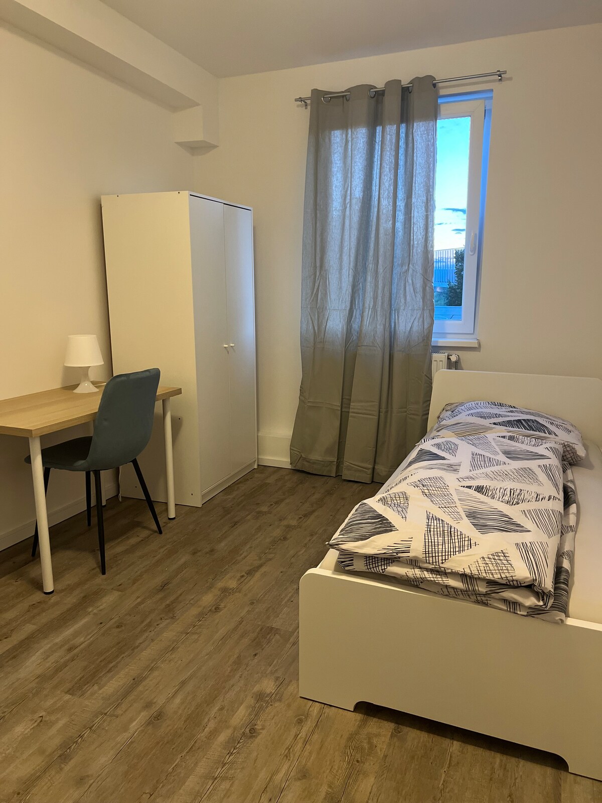 A small bedroom is presented with a single bed dressed in patterned bedding. A desk with a lamp is positioned next to the bed, and a wardrobe stands nearby. Natural light enters through a window adorned with a light curtain, enhancing the room's functionality.