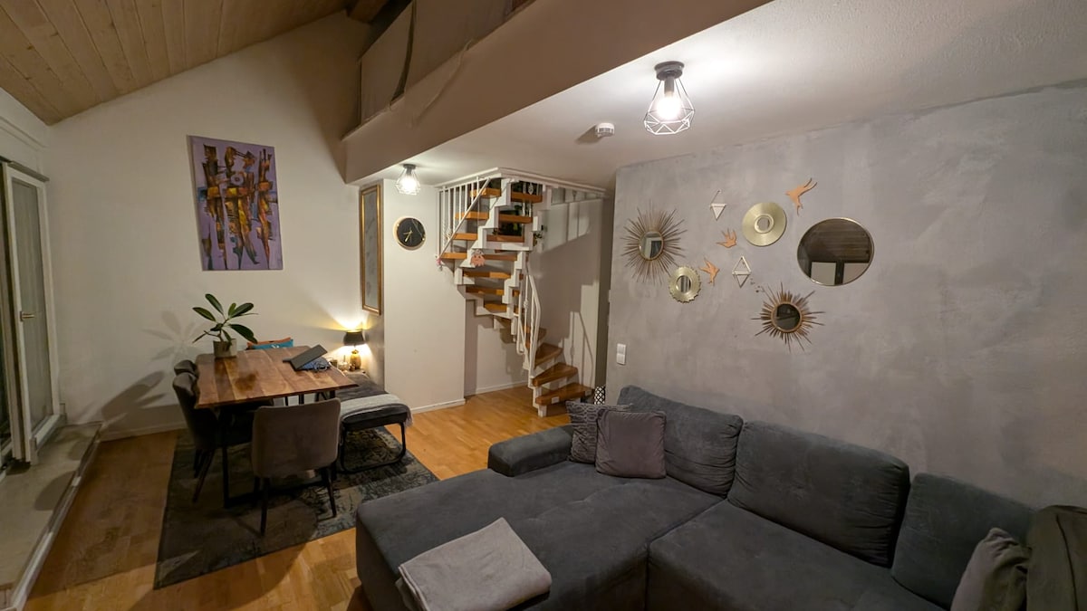 An inviting living space features a plush gray sofa with accent pillows. A dining table with four chairs stands nearby. Above, a staircase leads to an upper level. Wall art and decorative mirrors add visual interest, while warm lighting creates a cozy atmosphere.