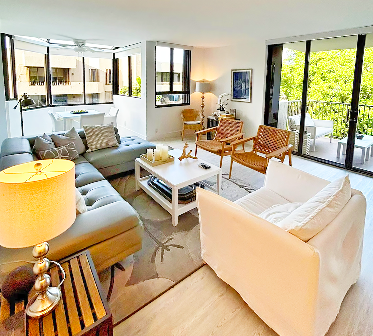 A spacious living area is highlighted by large windows that allow natural light to fill the room. Comfortable seating is provided by a sectional sofa and armchairs, complemented by a white coffee table. A lamp and side table add a subtle touch of warmth.