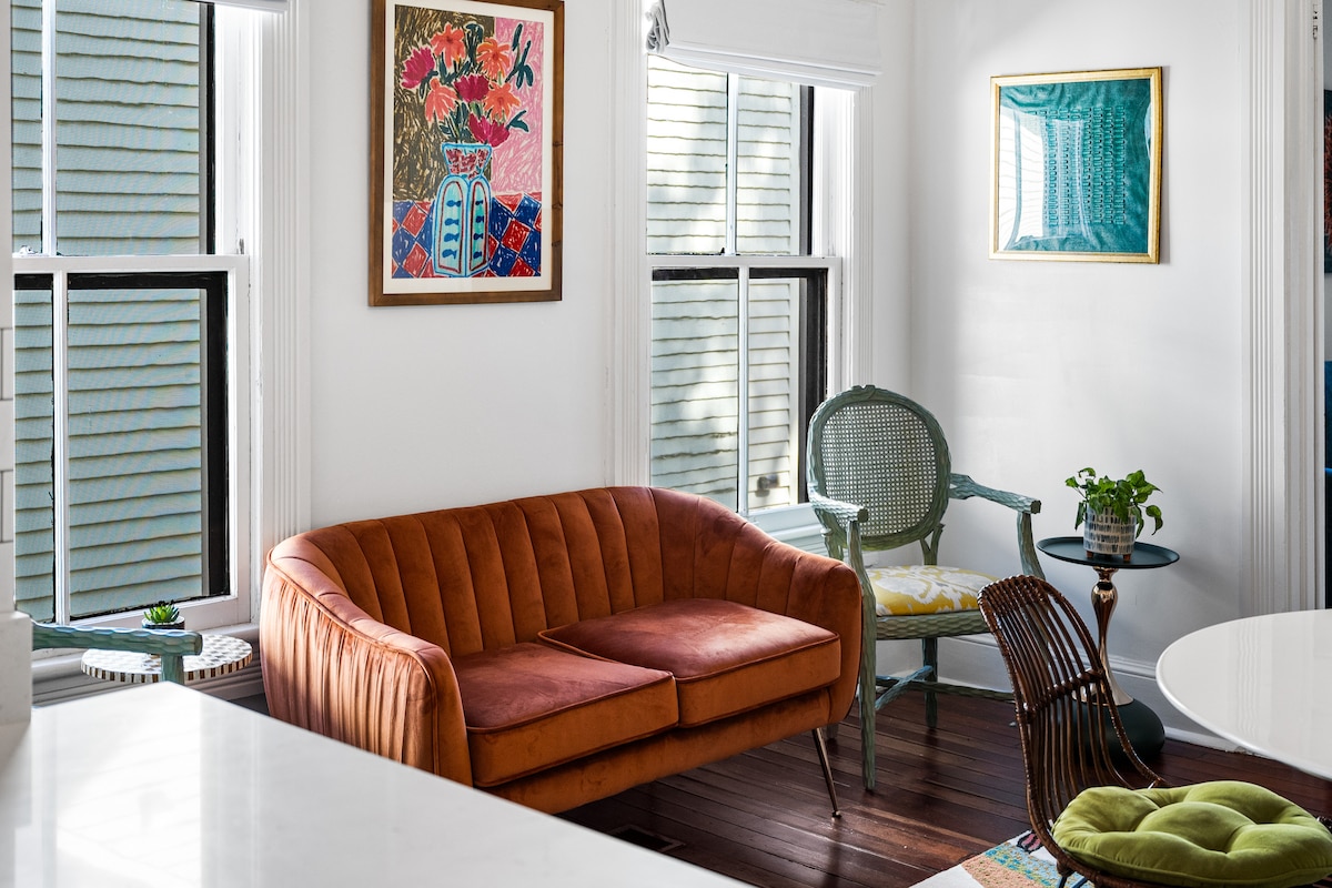 A cozy seating area is presented with a vibrant orange couch and an elegant green accent chair. Natural light fills the space through large windows, highlighting floral artwork on the walls. The hardwood floor adds warmth to the inviting atmosphere.