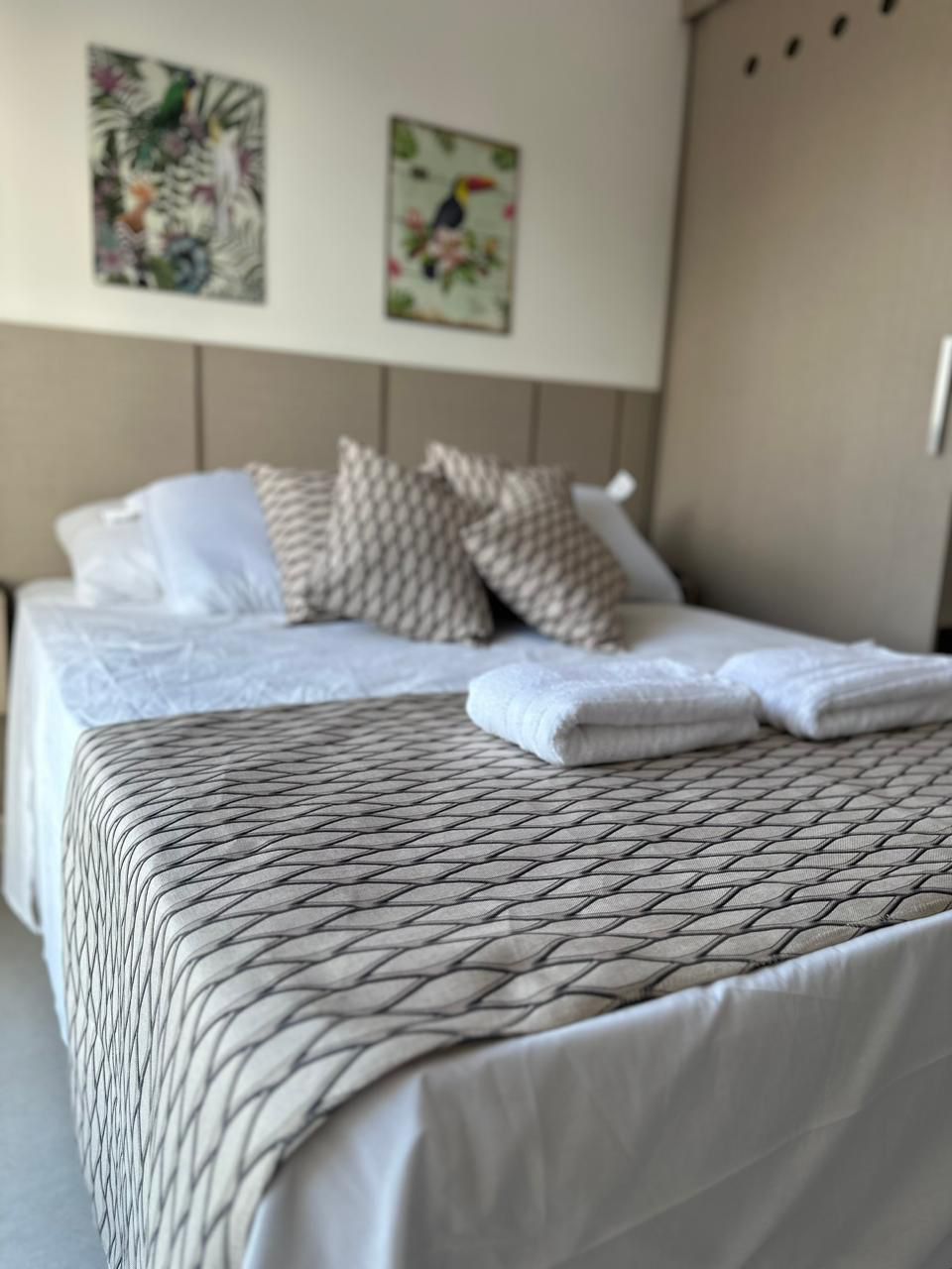 A spacious bed is positioned in a well-lit area, adorned with decorative pillows. Two art pieces featuring nature themes are displayed on the wall above the bed. Fresh linens and neatly rolled towels are presented at the foot of the bed, contributing to a tidy environment.