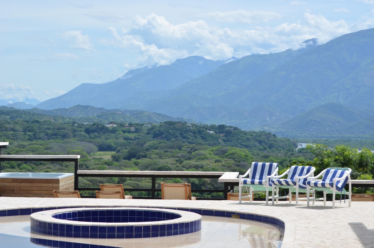 A view of the expansive natural landscape features rolling green hills and distant mountains under a clear sky. Lounge chairs with blue and white stripes are arranged around a circular hot tub, offering a serene outdoor setting for relaxation.