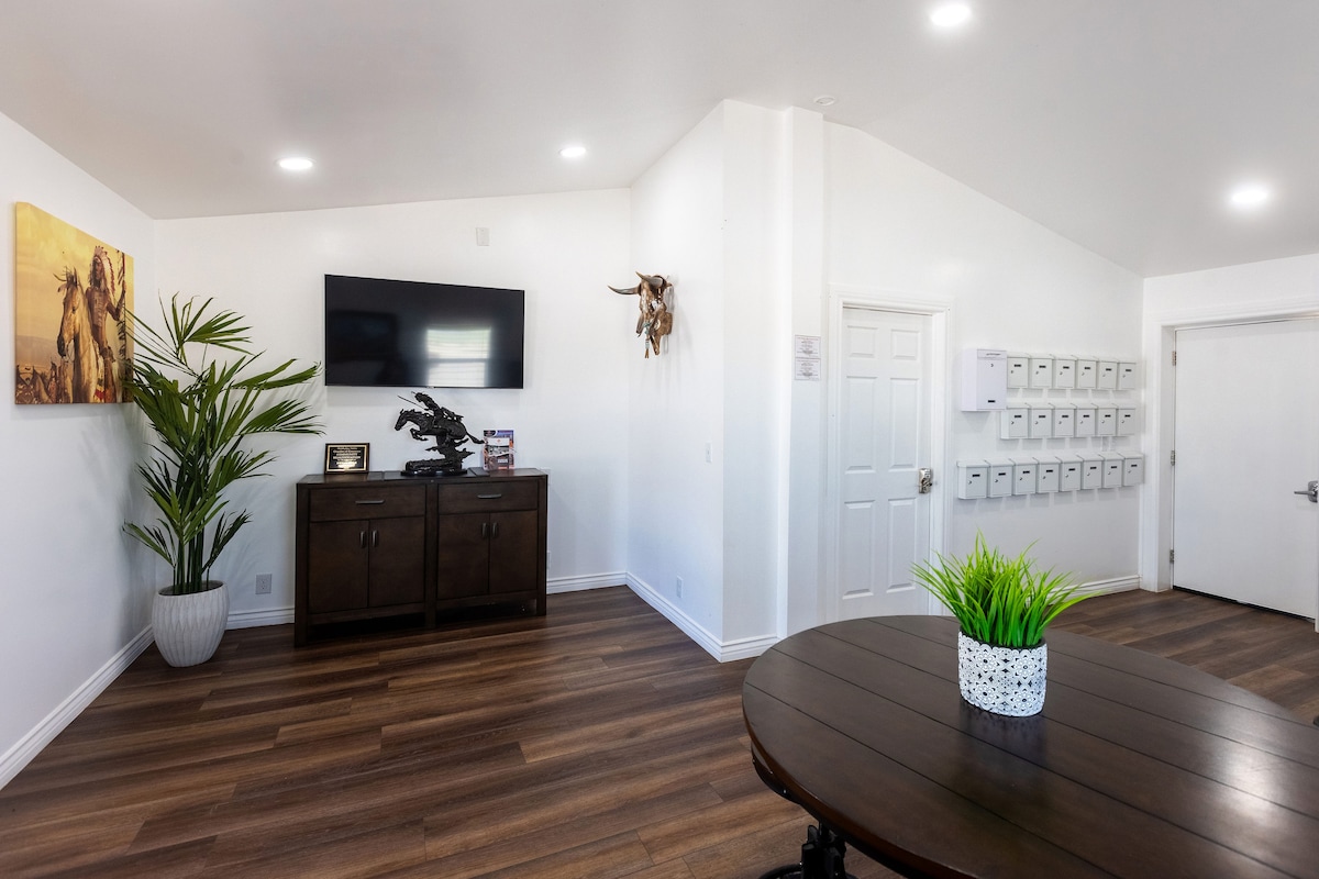 An inviting common area features a wooden table surrounded by a plant and a well-lit wall adorned with art. A dark wood cabinet holds a television, and a door leads to an exit. Mailboxes are mounted on the wall, adding to the functional design.