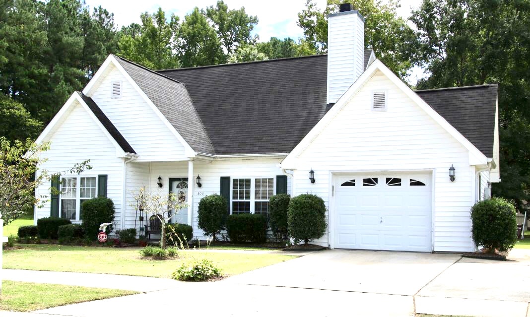 A single-story home is set amidst lush greenery, featuring a well-manicured lawn. The exterior showcases white siding and a black shingle roof. Large windows with shutters complement the inviting entrance, while a two-car garage and driveway provide convenient parking options.