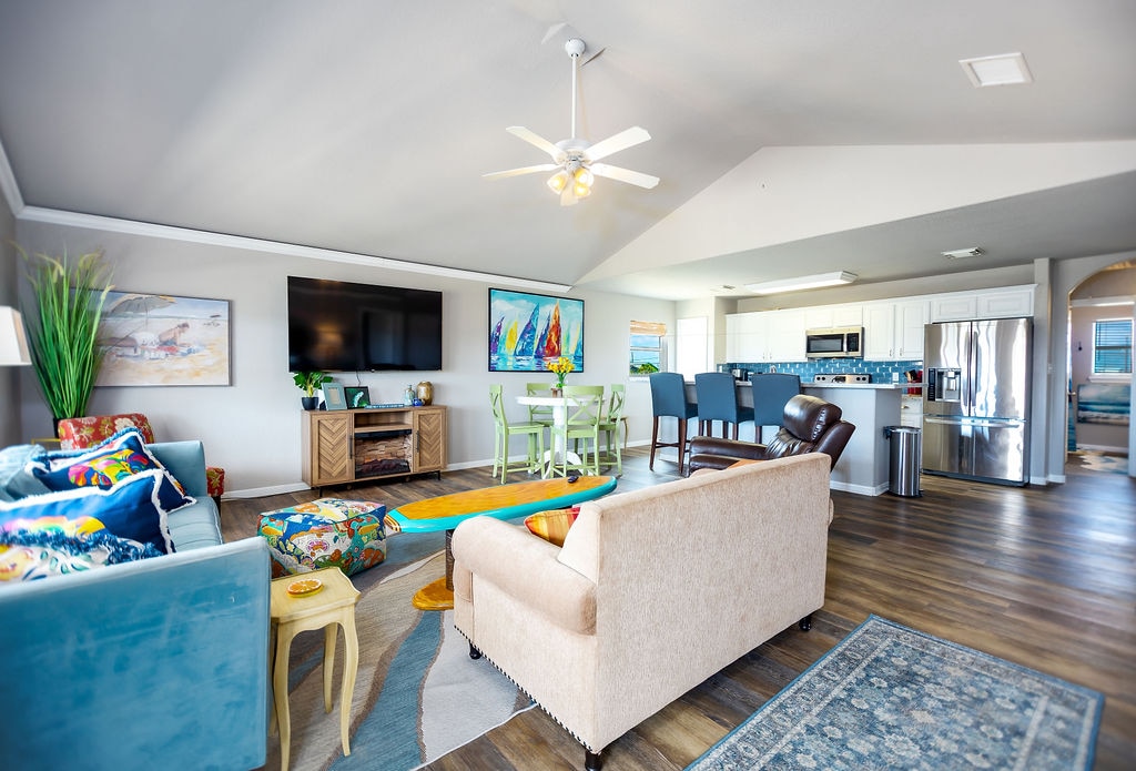 An open-concept living area features comfortable seating with colorful accents, a large wall-mounted TV, and a ceiling fan. A dining table and kitchen area are visible in the background, with natural light illuminating the space through multiple windows.