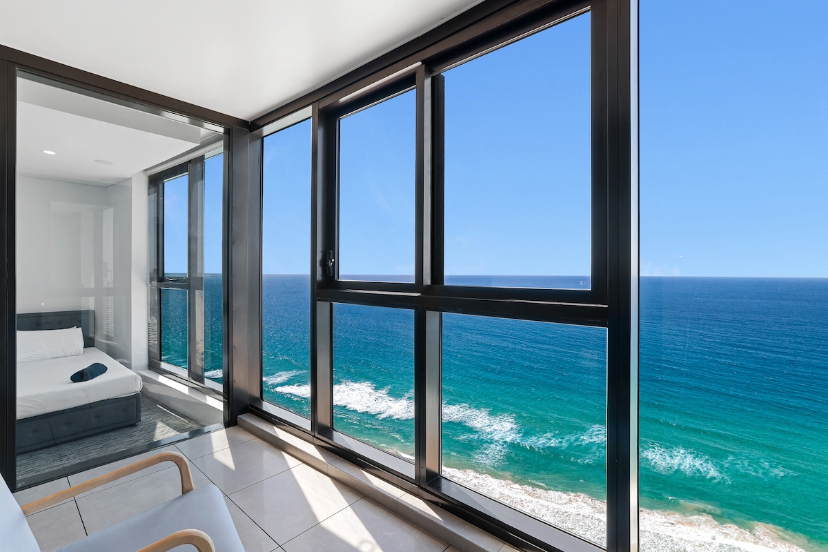 A bright living area overlooks the ocean through expansive floor-to-ceiling windows, creating a panoramic view of the coastline. The clear blue sky complements the tranquil sea. A cozy seating area is positioned near the windows, inviting relaxation while enjoying the scenery.