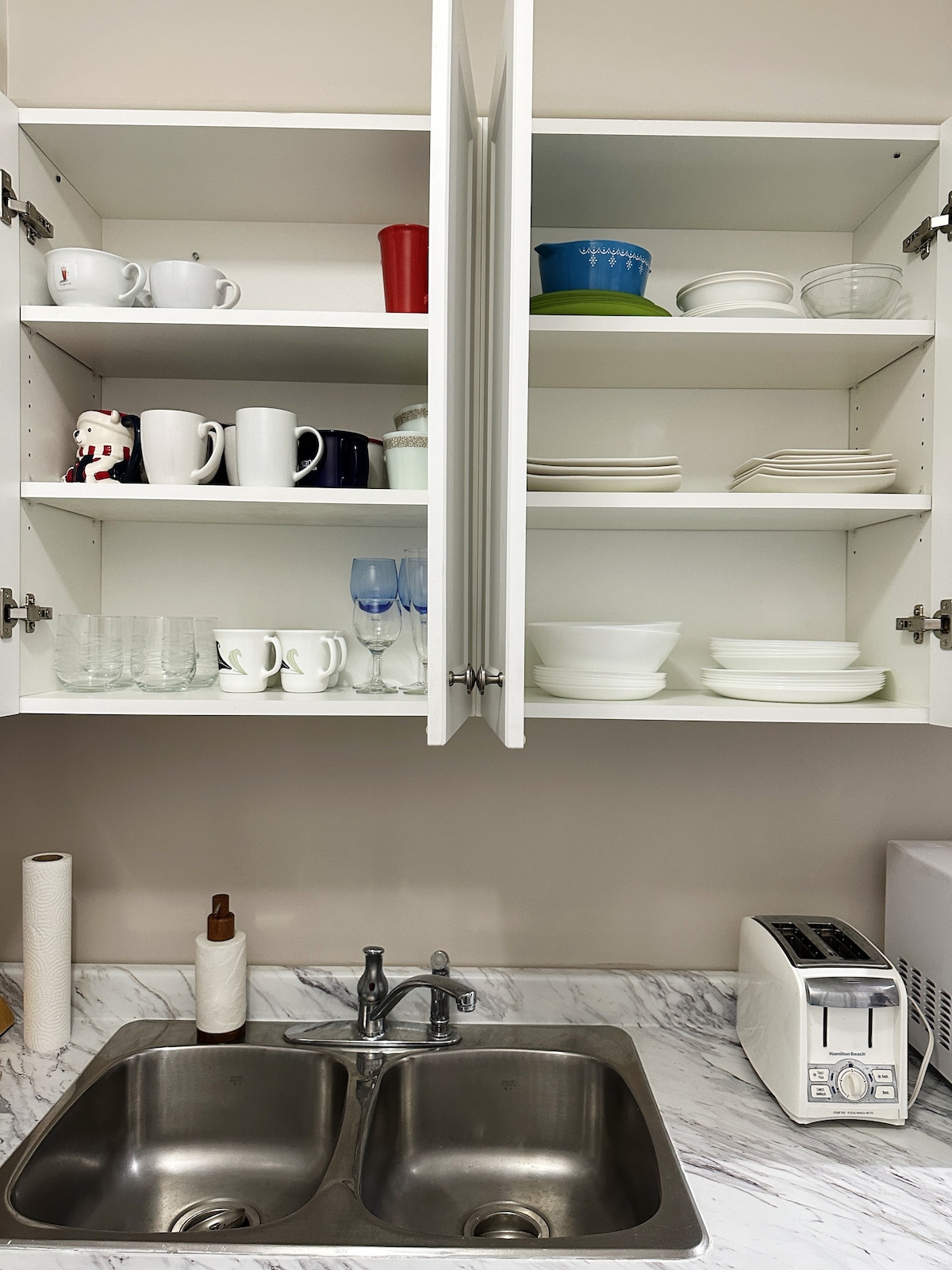 Kitchen cabinetry features an organized arrangement of clear, colored, and patterned dishware. Glassware and mugs are visible alongside stacks of plates. A double sink with silver faucets sits below, accompanied by a stainless steel toaster on the counter.