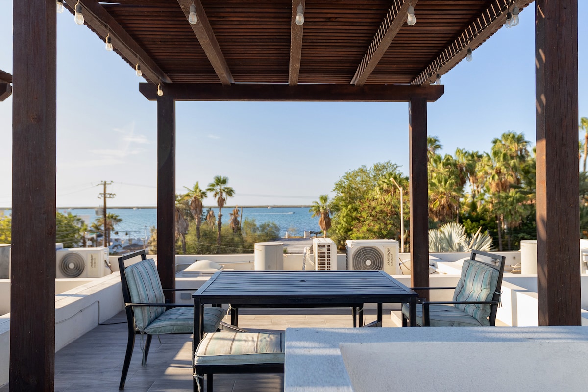 The rooftop terrace features a dining table surrounded by comfortable chairs, with a view of the nearby greenery and the sea in the background. Wooden beams support the structure, and string lights add a subtle ambiance against the clear sky.