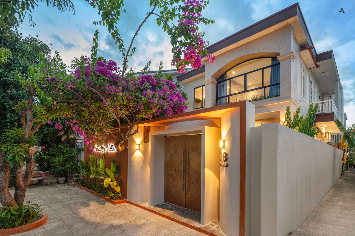 The exterior of the property features a modern facade with a welcoming entrance. Lush greenery and vibrant purple flowers surround the entrance, enhancing the tropical feel. Large windows reflect the sky, providing a glimpse into the inviting interior spaces.